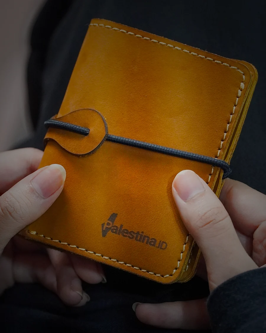A vegetable-tanned leather wallet held in two hands, saddle stitch and Palestina.id stamp in raking light